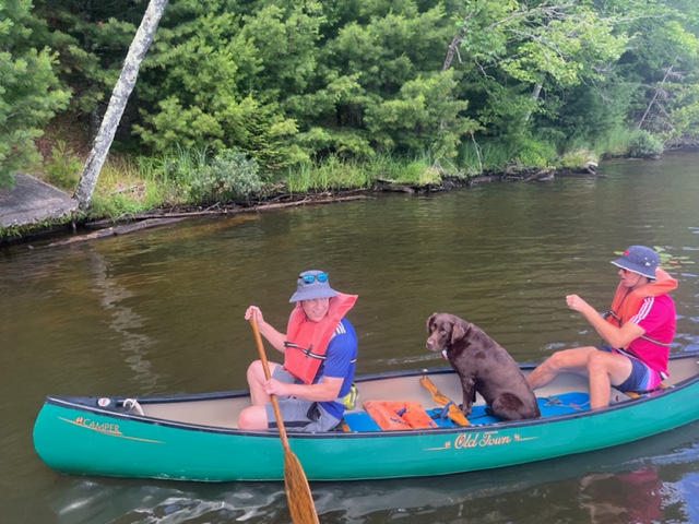 Photo of two men with hats canoeing with a dog sitting in the middle of the canoe.