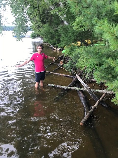 Photo of young man standing in the shoreline of the lake in front of the latticed logs.