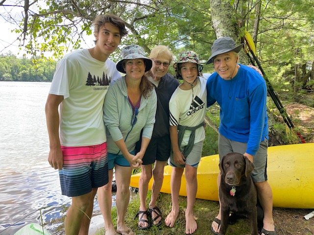 Five people posing for a picture on the shore of a lake with Rey, the chocolate lab.