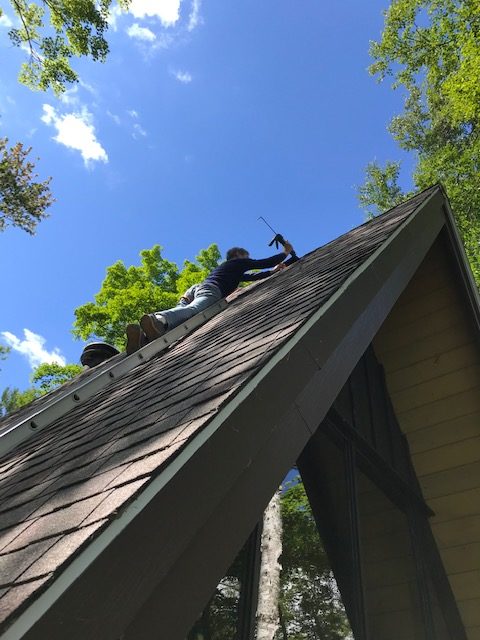 photo of man on a ladder fixing some shingles on the side of an A-frame roof.