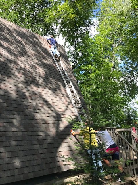 photo of man on a ladder fixing shingles near the top of an A-frame roof.  Two other men are holding the ladder at the bottom.