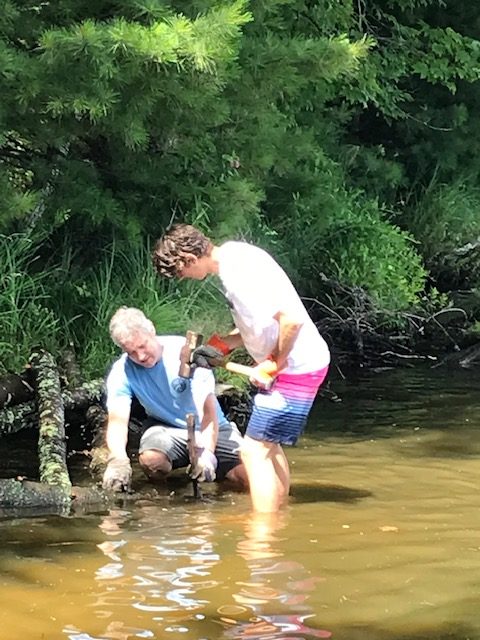 Photo of two men placing and hammering in logs along a lake shoreline.