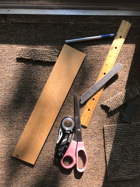 photo of tools to replace the carpet.  Includes the carpet, ruler, scissors, pen, razor blade, and a small wood plank.