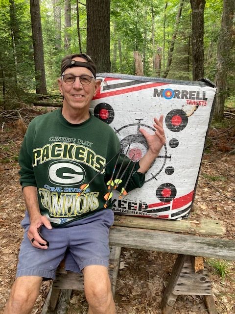 photo of man wearing a Green Bay Packers sweatshirt posing in front of an archery target and his arrow.